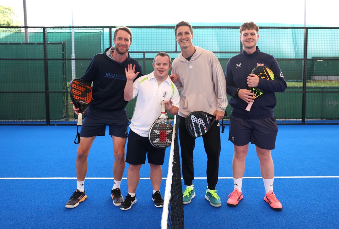 A group of four men stood on a blue sports court, either side of a net. They are all holding bats of a variety of colours.