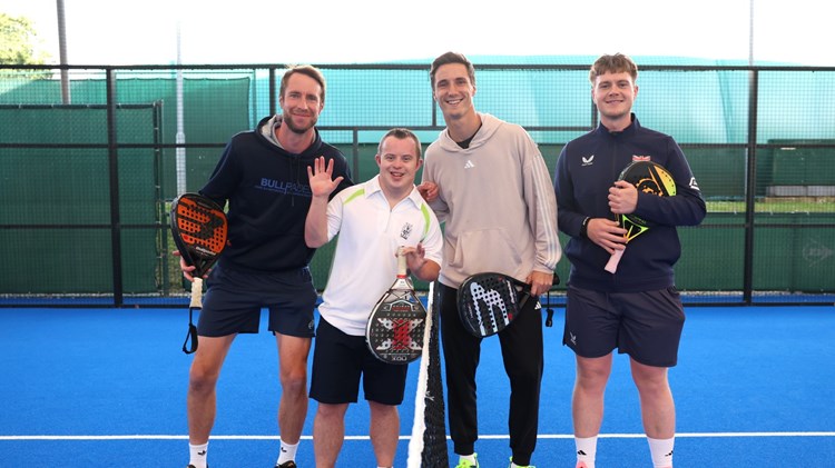 A group of four men stood on a blue sports court, either side of a net. They are all holding bats of a variety of colours.