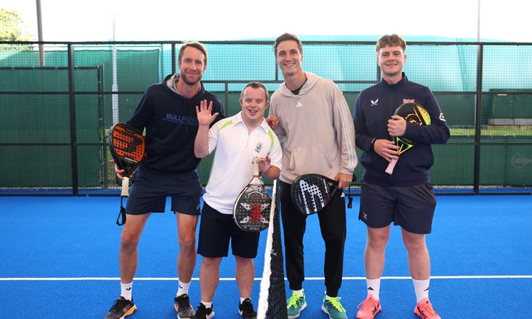 A group of four men stood on a blue sports court, either side of a net. They are all holding bats of a variety of colours.